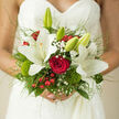 In this image, a hand-tied bridal bouquet rests against the finely detailed lace of a wedding gown, the filigree pattern of the fabric forming a romantic backdrop to the flowers. The overall palette is a graceful blend of creamy white, deep red, and rich green, composed with the kind of balance that comes from a florist who knows every petal by touch. At the heart of the bouquet sits a single, perfectly formed red rose, its velvety petals a deep, passionate crimson that draws the eye and anchors the design. Surrounding it, open white lilies and a few closed buds create a sense of movement and unfolding promise, their surfaces almost satiny where the light grazes them. Clusters of baby's breath gather like soft clouds, and glossy red berries nestle between the stems, bringing a playful, festive energy reminiscent of a winter wedding reception in a Clerkenwell warehouse venue near Farringdon Road. Mossy green accents and small tufts of dianthus introduce a fresh, garden-like quality, as if the bouquet carries a hint of Charterhouse Square's greenery into the celebration. Slender blades of grass and wispy foliage curve outward from the central blooms, framing the bouquet in a halo of gentle texture. The lighting is natural and flattering, making the whites glow softly against the gown while the reds deepen with richness. It feels like a bouquet designed for a bride walking through the cobbled streets around Clerkenwell Green, where historic churches and modern studios share the same skyline, a tangible expression of devotion, joy, and the quiet certainty of saying I do.