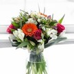 Evoking the vibrancy and creative edge of Clerkenwell, this cheerful floral arrangement bursts with life and colour. At the heart stands a luminous orange Gerbera daisy, its flawless petals and vivid centre instantly suggesting celebration and warmth-a floral sunbeam amid the urban energy of St John Street. Surrounding the gerbera, deep magenta and crimson roses, some fully opened in lush profusion and others as tightly spun buds, offer a rich romantic counterpoint. White alstroemeria with their distinctive striping, alongside clusters of small, daisy-like chrysanthemums, gently weave through the arrangement, adding layers of lightness and intricate texture. Creamy white and pale yellow roses bring a note of calm softness above, while flashes of lavender and purple blooms deepen the palette, echoing the creative flair found in Clerkenwell's galleries and studios. Emerald-green foliage envelopes the bouquet, from its sweeping, broad leaves cradling the base to the elegant, upright greens that sculpt the shape for a striking modern feel. The entire display sits within a minimalist glass vase, allowing the clarity of each submerged stem to shine through crystal water. Set against bright, diffused daylight pouring in-akin to the atmosphere of a Clerkenwell café on a spring morning-the bouquet exudes freshness and welcoming warmth. This is a floral arrangement that feels perfect for birthdays, congratulations, or simply to brighten someone's day in EC1, channeling both contemporary style and heartfelt emotion.