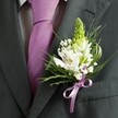 A close-up, high-angle photograph captures the upper torso of a man dressed for a formal celebration in Clerkenwell, with the camera lingering on the left lapel of his charcoal grey suit jacket. The fabric is smooth and finely tailored, the shoulders cleanly structured, and beneath the open lapel you can see a crisp, pale dress shirt with a faint grid pattern, neatly framed by a solid lavender tie that falls straight and precise down the centre of his chest. Pinned to the lapel is a carefully crafted boutonniere, the true focal point of the image, glowing softly as though lit by muted daylight from a nearby London window. At the top of the buttonhole stands a slender, pale green spire of tightly packed buds, perhaps hyacinth or Muscari, giving a sense of upward movement and new beginnings. Around this vertical accent, several open white Alstroemeria blossoms unfurl, their petals touched with fine streaks of burgundy and hints of golden stamens at their centre. Fine clouds of white gypsophila, or baby's breath, weave between the larger blooms, bringing a delicate, almost mist-like softness. Lush green foliage frames the flowers: thin, grass-like blades gently curve along the edge, while darker leaves lend depth and contrast to the white and lavender palette. At the base, a small satin bow in matching lavender ribbon encircles the stems, echoing the groom's tie and creating a harmonious, coordinated look. The overall feeling is one of quiet sophistication, the sort of refined buttonhole you might wear to a Clerkenwell registry office wedding or a chic city reception, where every floral detail has been thoughtfully composed to complement the suit and the significance of the day.