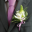 In this detailed close-up image, the viewer is drawn into the world of formalwear and fine floristry, centred on a single lapel adorned for a significant celebration. The wearer's dark charcoal grey suit jacket is tailored with clean lines and a sharply pressed lapel, resting over a white dress shirt that carries a very fine dark pattern, almost like miniature checks glimpsed between fabric folds. A sleek lavender necktie runs down the middle of the frame, its classic knot sitting snugly at the collar and the silky surface reflecting a subtle, elegant shine. Nestled against the lapel is a meticulously constructed boutonniere featuring white alstroemeria, or Peruvian lilies, their petals slightly cupped and painted with slender reddish-brown streaks and a touch of golden yellow near the centre where stamens peek through. Standing proudly at the heart of the design, a tall conical green bud provides an unexpected sculptural element, its fresh lime-green tone enlivening the composition. Tiny white clusters of baby's breath hover between the blooms, adding a light, frothy contrast. Around them, layers of greenery are artfully placed: thin, spiky grasses, feathery fern-like textures, and broader deep green leaves that create a lush, well-balanced frame. A narrow lavender satin ribbon, perfectly colour-matched to the tie, is wrapped and tied into a tidy bow at the base, anchoring the flowers to the lapel. The overall atmosphere suggests a moment of calm just before an important event, perhaps a wedding reception in one of Clerkenwell's converted warehouses or a prom gathering near Exmouth Market, where the wearer is about to step into an evening of photographs, toasts, and shared stories.