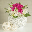 A rounded white wicker basket holds an intimate cluster of fuchsia blooms and ivory alstroemeria, the composition lit by a soft, diffused glow that feels like late afternoon light filtering through Clerkenwell loft windows. At the centre, the fuchsia flowers are plush and generous, their petals folding over one another in ruffled layers, creating depth and richness reminiscent of romantic bridal posies. Surrounding them, creamy alstroemeria open in star-shaped whorls, each flower marked with fine brushstrokes of pink and soft gold, so detailed they appear hand-painted. The interplay between the two colours creates a sense of quiet luxury rather than loud contrast, like the polished yet understated style often seen in weddings around Charterhouse Square. Slender green stems and leaves thread through the arrangement, some tipped with tight buds that promise more blooms to come, lending a sense of growth and fresh beginnings that suits engagement parties and registry weddings nearby. The white wicker basket is tightly and evenly woven into a rounded, almost sculptural form, its matte surface catching the light along each curve and echoing the clean lines of contemporary Clerkenwell interiors. On the tabletop, two pale cream wicker spheres rest casually beside the basket, their open latticework and gentle shadows adding dimension and visual rhythm, as if part of a modern wedding tablescape. The background is pale and uncluttered, allowing the flowers' colours and textures to stand fully in focus, like a centrepiece displayed on a simple white cloth in a local town hall. This arrangement would be ideal as a bridal gift or small table design for intimate celebrations around Farringdon, with every petal suggesting care, romance, and the freshness of newly made promises.