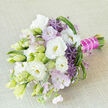 A sophisticated hand-tied bouquet rests at a diagonal across a pale beige, lightly textured fabric, the neutral backdrop allowing its whites, purples, and greens to quietly glow. The composition is anchored by generous white lisianthus, some fully open with creamy, softly cupped centres, others still in elegant bud, their pale green casings promising future bloom. Between these, wisps of lavender-pink phlox and other pastel blossoms introduce an airy, gauze-like softness, as though mist drifting over Clerkenwell Green at first light. Spherical purple allium, made up of star-like florets, stand proud among the more delicate heads, adding sculptural drama and a deeper violet note to the palette. Around and beneath the flowers, fresh foliage in many shades of green gives depth and rhythm: slim leaves, curled loops of bear grass, and lighter spiky accents that echo wild meadow textures while remaining carefully controlled. The stems are gathered firmly into a neat bundle, the handle dressed in bold magenta satin. A line of tiny white pearls runs along the wrap, their subtle shine suggesting the polished details of a bespoke bridal design, perhaps destined for a ceremony in one of Clerkenwell's converted warehouses or historic townhouses. The bouquet feels fresh yet timeless, its style sitting somewhere between classic garden romance and modern floral artistry. Even without scent, the image suggests a gentle fragrance and the cool touch of just-cut stems, as if the flowers had been prepared only moments ago in a local studio near Farringdon Road, ready for personal delivery to mark a wedding, anniversary, or thoughtful celebration.