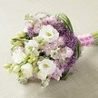 A beautifully rounded bridal bouquet lies serenely on a pale, textured fabric-it's a floral masterpiece woven with inspiration from Clerkenwell's winding lanes and creative flair. Tucked at its core, pillowy white lisianthus blossoms glow softly, unfolding their layered petals around delicate yellow centers. Sprigs of pale pink and lavender stock flowers provide ruffled, romantic accents, echoing the playful energy of Exmouth Market. Arcing through the gentle blooms, deep purple allium 'Star of Persia' bursts add a bold dash of colour and sculptural intrigue, calling to mind the striking contrasts seen in Clerkenwell's mix of old and new. Lively, fresh green foliage, slender and youthful, traverses the bouquet-each leaf and bud infusing the arrangement with natural vitality and textural depth. The flowers are bound at the base with radiant fuchsia satin ribbon, wrapped with a band of white pearls for a subtle hint of bridal luxury and joy. Bathed in natural, even light, the bouquet's intricate hues and tactile textures come alive, radiating a sense of romance and celebration. Crafted with care by local artisans, this bouquet would be equally at home as a showpiece at a Hatton Garden wedding, a statement for a summer ceremony in Spa Fields, or a thoughtful gift for someone cherishing those first moments of commitment in Clerkenwell.