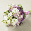 A carefully hand-tied bouquet lies horizontally on a pale beige, finely textured fabric that could be linen or softly woven jute, its neutral backdrop echoing the warm stone and brickwork often seen around Clerkenwell's historic streets. The eye is first drawn to the large white lisianthus blooms, their petals layered and velvety, opening generously to expose subtle yellow-green centres while neighbouring buds remain gently furled, suggesting a moment captured between promise and full blossom. Encircling these focal flowers, clusters of soft pink to light mauve stock provide a dreamy haze of colour, each stem filled with many small, ruffled florets that lend the bouquet a romantic, almost cloudlike fullness. Punctuating this softness are clusters of spiky, starburst purple filler flowers, similar to ornamental allium, whose pointed petals radiate outwards and introduce lively texture and a vivid lavender-purple note. Long, supple blades of deep green foliage arc and loop through the composition, particularly along the right edge, where their graceful curves guide the gaze and add structure without disturbing the bouquet's gentle character. The stems are neatly aligned and cinched with a bold fuchsia satin ribbon, its rich sheen catching the light, while a fine strand of tiny white pearls drapes over the wrap, adding a hint of bridal jewellery elegance. The scene feels calm yet celebratory, like a bouquet set down for a moment on a windowsill near Clerkenwell Green during a wedding, anniversary, or intimate gathering, radiating expert design, tenderness, and a sense of cherished occasion.