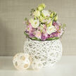 On a smooth white surface reminiscent of a bright Clerkenwell loft kitchen counter, a lush, rounded floral arrangement commands attention without any sense of fuss. The flowers spill gracefully from a striking spherical vase crafted from interlaced white twigs, forming an airy woven orb that reveals stems and shadows within. Nestled inside this sculptural container, large lisianthus blooms in soft cream and buttery ivory unfold in many layers, their velvety petals catching the light. Around them cluster delicate pink stock flowers, their ruffled florets stacked along slender stems, adding both fragrance and a dreamy, romantic softness. Small, star-like purple allium heads peek between the pastels, bringing a playful pop of colour and a subtle architectural edge to the design. Fresh greenery weaves through the bouquet, with darker green leaves neatly wrapping the lower portion of the arrangement to give a clean, cinched finish, almost like a natural ribbon. To the left, two decorative wicker spheres made from pale, natural wood strips sit on the tabletop, one slightly larger than the other; their open basket weave mirrors the lattice pattern of the vase and adds a relaxed, organic rhythm to the scene. The background is a softly blurred neutral wall in tones of beige and greige, textured enough to suggest fabric or grasscloth but understated enough to let the flowers glow. The overall impression is one of tranquil elegance-a piece that would be perfectly at home in a Clerkenwell design studio, a converted warehouse apartment near Exmouth Market, or a quiet reading nook. It feels ideal for thoughtful gifts, intimate celebrations, or simply bringing gentle colour and calm to everyday city life.