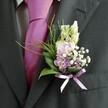 A detailed close-up shows the lapel of a sharply tailored charcoal suit, the fabric subtly textured and softened by natural daylight, as if the wearer is standing just outside a Clerkenwell registry office or near Exmouth Market before the ceremony begins. The crisp, pale dress shirt beneath carries a gentle grid pattern that adds quiet structure, while a vivid magenta silk tie, flawlessly knotted, introduces a confident flash of colour. Pinned neatly to the left lapel is a meticulously crafted wedding buttonhole, its floral artistry stealing the scene. At the top rise slender, unopened buds of creamy white or pale vanilla, reminiscent of snapdragons or lisianthus, their smooth, elongated forms suggesting promise and patience. Nestled below are clusters of soft pink and delicate lavender blooms-sweet-pea-like petals or miniature stocks and orchids-that gather together in a cloud of gentle colour. Interwoven sprigs of white gypsophila create a fine, misty veil, while a few deeper purple florets add nuance and depth, much like the shaded corners of Spa Fields on a spring afternoon. The greenery is thoughtfully layered: broad, glossy dark green leaves form a protective cradle at the base, while feathery bright green foliage, similar to asparagus fern, arcs outward in light, airy gestures. A satin ribbon in tender lilac is wrapped and tied in a precise little bow at the stems, its soft sheen echoing the purple tones above. The overall impression is of refined yet playful elegance, the sort of buttonhole a groom or treasured guest might choose for a modern London wedding in Clerkenwell, where tradition, style, and heartfelt celebration meet on the same cobbled streets.