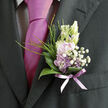Pinned against the rich, dark weave of a formal suit, this lilac and white boutonnière appears as a soft breath of spring drifting into a Clerkenwell celebration. At its heart, pale lavender blossoms open in delicate layers, each petal slightly translucent, catching and softening the ambient light like stained glass in a nearby Farringdon church. Nestled beside them, creamy white buds and tiny white filler flowers create a gentle contrast, their rounded forms and frothy texture suggesting early blossom along the tree-lined paths towards Gray's Inn Road. A collar of smooth, glossy green leaves cradles the miniature cluster, their curved edges wrapping partway around the blooms to give a feeling of shelter and composure. Slender, grass-like stems and airy sprigs of greenery emerge behind, adding movement and giving the piece a subtly untamed edge, as though the flowers were freshly picked from a London garden just before the ceremony. The stems are bound carefully with a sheer lavender ribbon, tied snugly so that only a neat, tapered bundle peeks from the lapel, mirroring the nearby tie in both tone and quiet elegance. You can imagine the soft, sweet fragrance of the flowers mixing with the crisp scent of pressed fabric and polished shoes as guests arrive. The overall impression is one of refined intimacy: a meticulously crafted detail that speaks of romance, thoughtfulness, and the steady hands of an experienced florist used to preparing wedding flowers for venues across Clerkenwell and the surrounding streets.