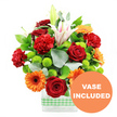 A joyful, contemporary bouquet overflows from a clear, square-edged glass vase, which is neatly wrapped around the base with a light green and white gingham ribbon that suggests relaxed gatherings and weekend brunches in Clerkenwell. The arrangement is tightly composed and visually abundant, starting with deep red roses whose velvety petals curl inwards to form rich, romantic blooms at the centre. Nestled among them are red-orange carnations, their ruffled petals creating soft waves of colour and giving the heart of the bouquet a plush, almost cloud-like texture. Between these, bold orange gerbera daisies with striking dark centres stretch their flat, daisy-shaped heads outward, echoing the warm, energetic feel of nearby Exmouth Market on a sunny afternoon. Pockets of bright lime-green button chrysanthemums are scattered throughout, their neatly rounded forms adding a playful pop of freshness and a modern, graphic edge. Rising above, white Oriental lilies streaked with pale pink lift gracefully from the mass of colour; some lilies are wide open, revealing delicate stamens, while others stay cocooned in pale buds, promising continued interest over the coming days. The bouquet is laced with deep green leaves and fine sprigs of silvery eucalyptus, giving depth, movement, and a gentle, aromatic quality that feels like stepping out into a leafy Clerkenwell side street after rain. The flowers are photographed against a pure white background, so every hue stands crisp and clear. In the lower right corner, a bold circular orange badge overlaps the glass, carrying the words VASE INCLUDED in clean white capitals, making it visually clear that the vase shown is part of the finished, ready-to-place arrangement.