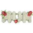 A dignified three-dimensional tribute reads MOM in large capital letters, each letter formed from hundreds of small, pristine white spray chrysanthemums that create a soft, cloud-like surface. The chrysanthemums are densely clustered to give a cushiony, voluminous texture; their petals suggest a quiet whiteness that conveys purity and peacefulness under soft, even daylight. Supporting the blooms is a subtle dark-green framework that peeks out along the letter edges, giving the shapes a clear structure while remaining unobtrusive. To punctuate the serenity, three glossy red anthuriums with their distinctive pale yellow-white spadices are placed as focal points: one on the upper left of the first M, one at the base of the central O, and one on the upper right of the second M. Each anthurium is surrounded by varied dark green foliage-feathery fern fronds and broader glossy leaves-that deepen the colour palette and add tactile contrast against the matte chrysanthemums. Thin arching blades of grass or slender stems sweep between the red blooms, visually tying the letters together and adding a gentle sense of movement across the composition. The overall effect is classic and comforting, an arrangement suited to a funeral, wake, or graveside service that honours a mother's memory with dignity and tenderness. The piece reads as a calm, crafted expression of love and remembrance, the white blooms offering solemn comfort while the red hearts of anthurium speak quietly of enduring affection and respect.