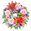 A large, perfectly round sympathy wreath is shown laid flat on a crisp white background, allowing every flower and leaf to stand out clearly. The ring is densely packed, beginning with a deep, textural layer of foliage: silvery eucalyptus, fine ferns, and varying shades of green that create a soft, natural frame. Nestled into this greenery are clusters of full, open roses in blush and lavender-pink, their many petals gently unfurled to give a feeling of tenderness and calm. Between the roses sit ruffled lisianthus, some a pale, almost cloud-like pink and others a rich, respectful purple, adding contrast and movement. Two striking coral-orange Asiatic lilies, one set into the upper part of the circle and one nearer the bottom, act as warm focal points, their star-shaped blooms and prominent stamens lending quiet drama and hope. Several tight lily buds emerge from the foliage, suggesting new life and the unfolding of memories over time. Spiky, steel-blue eryngium thistles bring a unique, architectural texture, while sprays of tiny lavender-blue statice weave through the design like a soft haze. Around the feature lilies, carefully folded, bright green leaves introduce a contemporary, considered touch that reflects the skill of a professional florist. This wreath, with its balanced mix of pastel comfort and subtle vibrancy, would sit beautifully at a funeral or remembrance gathering in Clerkenwell, perhaps in one of the area's historic churches or crematoria, expressing sympathy, respect, and unbroken love in a gentle, floral language.