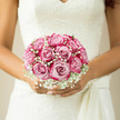 Cradled tenderly in front of her lace wedding dress, the bride's bouquet captures a moment of gentle calm amid the buzz of Clerkenwell streets. A rounded cluster of blush pink roses forms the heart of the design, each bloom perfectly shaped, their layered petals unfurling like soft silk in slow motion. Between them, airy clouds of white gypsophila thread delicately through the arrangement, softening every line and giving the bouquet a romantic, almost dreamlike haze. The interplay between the velvety rose petals and the fine, feathery texture of the baby's breath creates a tactile contrast you can almost feel, like tracing the edge of historic stone and new glass along Clerkenwell Road. Natural daylight spills over the flowers, highlighting gentle shifts in colour-from pale, nearly ivory pink at the edges to a deeper, warm blush toward each rose's centre-and casting faint shadows on the bride's intricately patterned lace. The stems are gathered neatly, wrapped with a smooth ribbon that disappears into her hands, suggesting poise, care, and craft. Imagining the fragrance, there is the soft sweetness of fresh roses, clean and romantic, just as you might notice when passing a florist's doorway near Farringdon Station. The bouquet's shape is balanced and symmetrical, neither too large nor too small, creating a sense of quiet confidence as the bride prepares to walk toward her ceremony. It feels timeless yet personal, the kind of classic design a local florist might create for a couple who cherish understated elegance and heartfelt emotion on their wedding day.