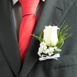 In soft ambient light the ivory rose of this boutonnière stands out with poised confidence against a smart charcoal jacket, the velvety petals layered into a compact, classic bloom that reads as both romantic and resolute. At the rose's base, a spray of baby's breath gathers like a whisper of cloud, its tiny white beads lending a sense of purity and lightness that tempers the rose's quiet strength. The composition is given further contrast by a checked shirt peeking beneath the lapel and the bold accent of a red silk tie nearby, which frames the boutonnière and makes its cool tones sing. Slender fern slivers and twisted blades of deep green arch gracefully around the central blossom, introducing movement and depth so the piece reads three-dimensionally when viewed from the side. A neat white ribbon bow at the stem finishes the work with a touch of ceremony; its smooth texture and soft sheen mirror the tactile freshness of morning-cut stems. The scent is subtle - a faint, rosy perfume that conjures dew on petals and the calm of a Clerkenwell morning, perhaps after a browse of Leather Lane market. As assembled by a practiced hand in our neighbourhood, this boutonnière is understated elegance for a groom or honoured guest who wants a refined, tactile emblem of commitment and celebration rather than ostentation. It is finished with careful wiring and tape so it sits securely on the lapel, moving confidently through vows, receptions or formal photographs without losing its composed silhouette.