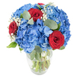 A cool, celebratory bouquet sits in a simple clear glass vase, its curved base and tapered rim revealing the green stems and water that sustain the blooms. At the heart of the arrangement are large, cloud-like clusters of sky-blue hydrangeas - each head built from hundreds of delicate, overlapping petals that catch the light and read almost like a summer sky after rain. Interwoven among the blue are three velvety red roses, their tight spirals of petals presenting a concentrated, passionate punctuation against the cool hydrangea mass. Tiny sprays of white baby's breath float between the bigger flowers like blown cotton, softening edges and lending a luminous, airy texture. Muted, rounded eucalyptus leaves and fresh green foliage thread through the composition, their silvery-green tones adding depth, a faint herbal perfume, and a pleasantly rounded silhouette. The bouquet is brightly lit from one side, which highlights the hydrangeas' subtle veining and the roses' satiny sheen while casting gentle shadows that enhance the three-dimensional form. The overall effect is both bold and comforting - a confident pairing of cobalt and crimson that feels at once modern and timeless. Envision this on a breakfast table in a Clerkenwell flat near Exmouth Market, or brightening a desk on St John's Square; its clean lines and fresh scent would lift a room without overwhelming it. Appropriate for anniversaries, a congratulatory gift, or to bring a strong, joyful accent into a home, the arrangement communicates warmth, celebration, and an attentive florist's hand in composition and proportion.