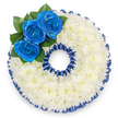 A bespoke funeral tribute shaped into a perfect numerical '0', captured against a clean white backdrop, made from a dense, cloud-like mass of tiny white spray chrysanthemums. From a close, top-down view you can see each florette packed shoulder-to-shoulder, creating a soft, pillowy texture that reads as both tactile and calming. A deep sapphire-blue border-pleated ribbon or dyed petals-traces the outer and inner edges of the zero, its ruffled sheen catching the light and giving the form a dignified, scalloped frame. At the upper-left curve, three royal blue roses are clustered with deliberate care; their petals open from tight centres into velvety spirals, the hue rich and saturated against the pure white chrysanthemum field. Glossy dark-green leaves tuck gently behind the roses, offering a fresh, natural counterpoint and guiding the eye around the circular negative space. There is a faint impression of scent-cool, green leaf notes beneath the subtle, powdery sweetness of chrysanthemum and the fuller, tea-like fragrance of the roses-yet the composition remains restrained and solemn. This personalised numeral tribute speaks of specific ages or meaningful numbers at memorials, wakes, or graveside services, where families in Clerkenwell and nearby Farringdon might choose it as a private expression of remembrance. As a local florist familiar with Exmouth Market and St John's Square, I select each chrysanthemum and ribbon fold for longevity, texture and the quiet dignity that such services require. The overall effect is one of ordered compassion: a pristine, contrasting arrangement whose cool whites and deep blues deliver a composed message of respect, comfort and lasting memory for those paying tribute.
