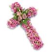 A deeply evocative floral cross arrangement is set against a luminous white background, its shape entirely defined by a dense covering of blossoms in soft to vibrant pink, mauve, and lilac tones. The cross has a plush, three-dimensional presence, with tightly clustered lisianthus flowers sweeping along each arm, their gently frilled petals overlapping like layers of fine fabric. The colour palette moves gracefully from very pale blush and cool lavender to richer rose-pinks, with many blooms showing darker, more intense centres that introduce visual depth. At the central intersection of the cross, three fully opened roses in a muted dusty mauve hue form an elegant focal group, their spiralling petals opening wide as if in a quiet gesture of farewell. Around these roses, sprays of fresh fern and bright green leaves weave through the flowers, offering contrast and a touch of garden-like freshness, reminiscent of the greenery you might find in a tucked-away Clerkenwell courtyard. Throughout the cross are tiny accent flowers in pink and soft purple, along with delicate green buds and scattered miniature white blooms, all contributing to a rich, intricate texture that rewards a lingering gaze. Along the outer contours, just visible beneath the thick carpet of petals, run slender ribbons or bands of darker purple, softly tracing the sacred form and lending a sense of ceremony. The arrangement feels calm and contemplative, like something lovingly prepared by Clerkenwell Flowers for a local funeral or chapel service, offering solace, spiritual comfort, and a beautiful visual symbol of remembrance in a time of loss.