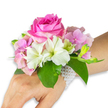 A delicate wrist corsage captured against a crisp white backdrop, arranged as a wearable whisper of spring. At its centre sits a full, deep fuchsia-pink rose whose tightly layered petals open in a soft spiral, their satiny texture catching gentle, diffuse light. Surrounding that rose, clusters of blush pink hydrangea florets form a lush, cloud-like cushion, each tiny petal offering a soft, almost translucent surface that contrasts with the rose's denser bloom. Several white alstroemeria (Peruvian lilies) fan out from the composition, their four-to-six petals tipped with pale green streaks near the throat, lending a fine, painterly detail. On one side a small grouping of pale green alstroemeria buds and a few barely open pink buds hint at future blossom and movement, introducing a tender sense of unfolding life. Broad emerald leaves cradle the bouquet, glossy and cool against the skin, setting off the pastel and vivid tones with striking contrast. The arrangement is secured to the wearer's left wrist by a refined multi-strand bracelet of small, luminous white pearls that wrap into a delicate net and trail a little along the back of a light-to-medium toned hand and forearm, visible here as part of the composition. There is a whisper of fragrance-fresh rose and clean green notes-suggesting this piece would be a serene companion at a wedding ceremony, prom first dance, or an intimate celebration. As a floralist might note while preparing pieces for clients around Clerkenwell's Exmouth Market or Spa Fields, the craftsmanship is careful and balanced: every stem is positioned for comfort, every leaf chosen to enhance the colour story, creating a timeless, wearable bouquet that reads as both elegant and personal.
