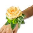 An elegant wrist corsage rests gently against a pale, neutral wrist, photographed on a crisp white background so every detail reads clearly for touch as well as sight. At the centre, a single large rose unfurls in soft peach-apricot tones, its layered petals milky and luminous like the inside of a shell; you can almost sense the velvety surface and the sweet, faint perfume that will lift in warm air. Nestled below the rose are two bright lime-green button chrysanthemums - tight, pom-pom spheres with a cheerful, tactile density that contrasts the rose's smooth curves and lends the design a modern, playful energy. A base of deep, glossy green leaves - the broad, supportive shapes of salal or ruscus - grounds the flowers and gives the wristlet depth and structure, while an artfully folded variegated leaf, striped with lighter green bands, sweeps around the bloom in a graphic arc that feels both architectural and soft. The corsage is posed on the inner wrist where it will be worn, the securing band hidden from view, allowing the arrangement to read as a seamless, wearable moment. Light falls softly across the petals and leaves, picking out textures and a gentle sheen on the foliage; the overall palette of peach, lime and rich green suggests celebration and quiet romance. Crafted with the care of a local florist who knows how a small piece like this can hold memory, it is perfectly suited for weddings, proms or anniversary evenings - an intimate floral accent that promises to brighten photographs and hands alike on a special Clerkenwell morning or festive evening.