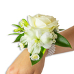 A refined wrist corsage photographed against a clean white backdrop, presented on a fair-skinned wrist that enters the frame from the lower right and tilts gently toward the center. At the heart of the design sits a creamy ivory rose, its velvety petals unfurling in a soft spiral that catches subdued, even light and gives the bloom a warm, pearlescent glow. Nestled just beneath and slightly to the left of the rose is a larger, trumpet-shaped white bloom-reminiscent of alstroemeria or a small lily-with a pale lime-green throat that draws the eye inward and adds a quiet depth of colour. Scattered through the cluster are several bright green buds, some tightly closed, promising future opening, and tiny sprays of delicate white gypsophila that float like breathy clouds around the main flowers, lending an airy, romantic texture. Lower on the wrist a cluster of small daisy-like white florets with sunny yellow centres gives a playful, fresh punctuation to the composition. Broad, glossy deep-green leaves frame the arrangement, their smooth, waxy surfaces providing a lush contrast to the soft white petals. The whole piece is anchored with a chic multi-strand pearl bracelet-uniform round pearls, cool and luminous against skin, looping comfortably and holding the flowers in place. I picture making this for a bride walking from St John Street to Clerkenwell Green or for a prom party leaving the Barbican, the scent faintly powdery and the ensemble quietly elegant-an understated accessory meant for vows, dances, and moments that ask for timeless refinement.