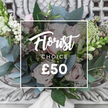 A hand-tied bouquet of soft, blush garden roses sits gently on an ornate, distressed cream console, the carved details of the wood blurred into the background so the flowers read as the quiet focus. The roses are petal-soft, their pale pink heads unfurling into velvet spirals that catch the light, while sprays of tiny white blooms-like waxflower or astrantia-drift through the composition to add an airy, cottage-garden feel. Silvery-blue eucalyptus leaves fan out, their rounded shapes cooling the palette and suggesting a subtle, calming scent that underpins the arrangement. Bright white berries punctuate the bouquet with little bursts of brightness, and deeper pink or burgundy buds tuck into shadowed corners to give depth and gentle contrast. Tall, slender green-and-white spikes peek at the edges, lending a vertical rhythm reminiscent of snapdragons and giving the silhouette a relaxed, garden-picked movement. At the stems a soft cream satin ribbon is tied in a simple, elegant knot, reinforcing the bespoke, hand-finished quality. The overall mood is rustic-chic and effortlessly romantic-perfect for birthdays, anniversaries, or as a spontaneous act of kindness-captured in soft daylight that enhances the natural hues and textures. This feels like a bouquet brought home from a morning walk past Leather Lane market and placed on the mantel at St John's Square, a thoughtful floral choice for someone who loves gentle colour, tactile contrast, and the calm confidence of skilled floral craft.